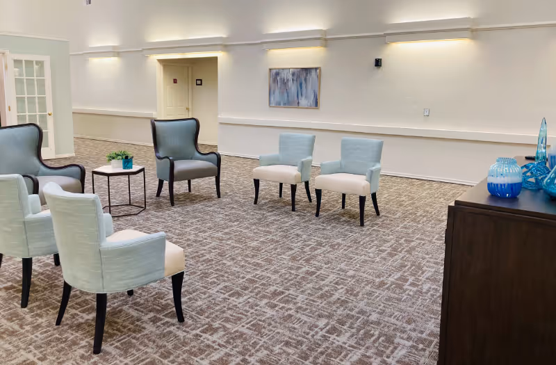 Bright, carpeted common room with light-blue and gray chairs arranged in a conversational seating area and wall art on the far wall.