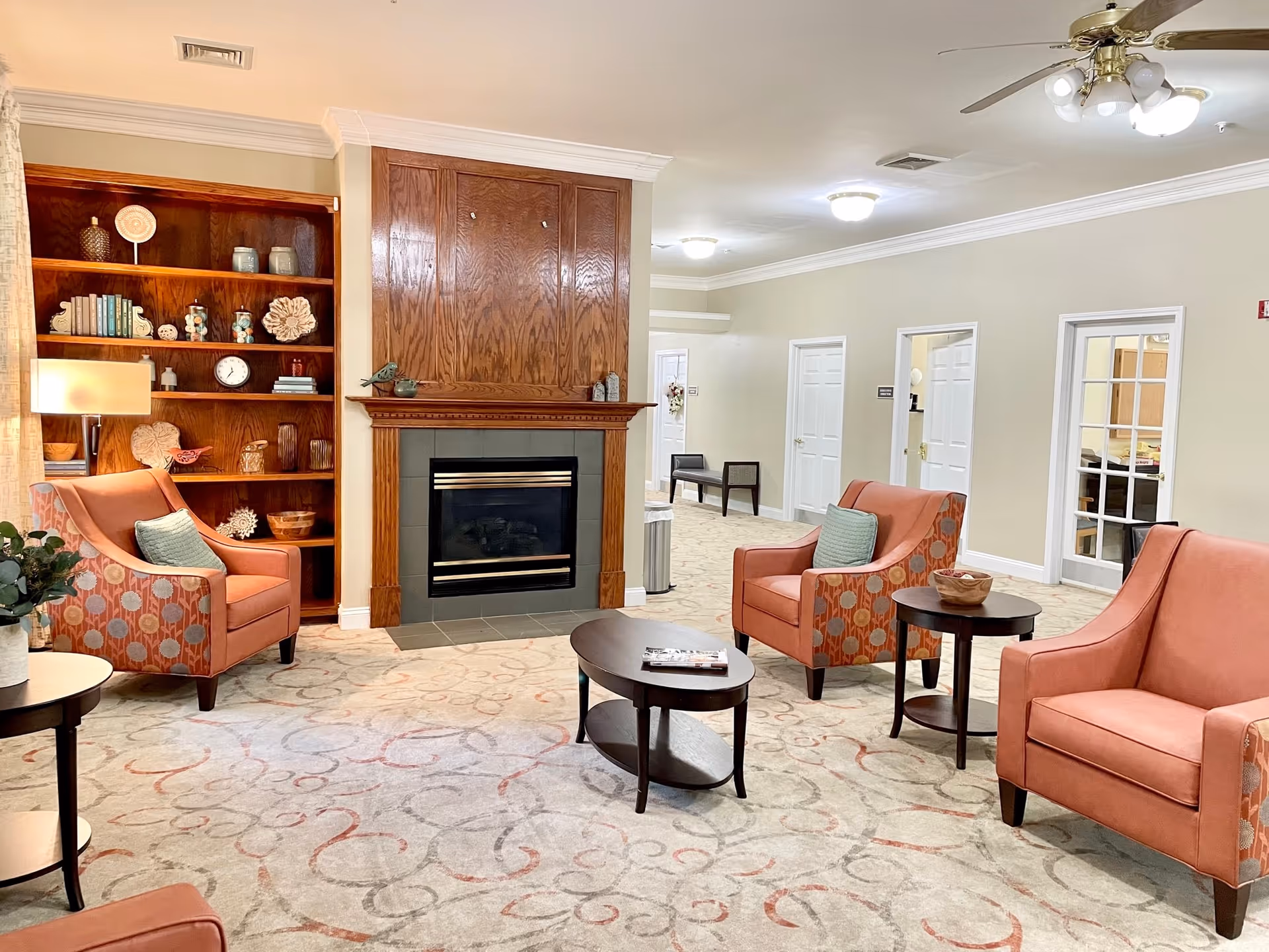 A cozy senior living facility common area with three coral-colored armchairs arranged around two dark wood coffee tables. There is a fireplace with a wooden mantel and built-in wooden shelves filled with decorative items and books. The room has beige walls, a patterned carpet, and ceiling lights including a ceiling fan with lights. Doors and a hallway are visible in the background.