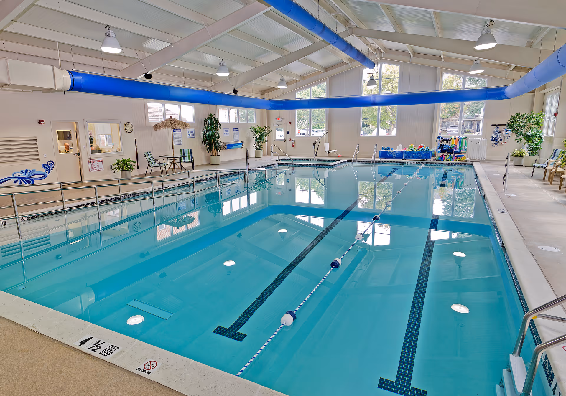 Indoor community swimming pool with marked lap lanes, seating, plants, and large windows.