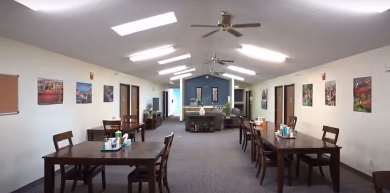 Interior view of a senior living facility common area with two rows of wooden tables and chairs on either side. The room has a high ceiling with ceiling fans and fluorescent lights. The walls are decorated with framed pictures, and there is a seating area with sofas and a fireplace at the far end.