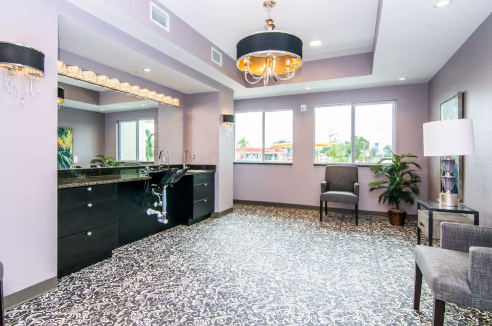 A spacious room with lavender walls and patterned flooring featuring a large mirror above a black countertop with two sinks. The room is decorated with modern light fixtures, a gray armchair, a potted plant, and a side table with a lamp. Large windows allow natural light to enter, showing an outdoor view with trees and a gas station in the distance.