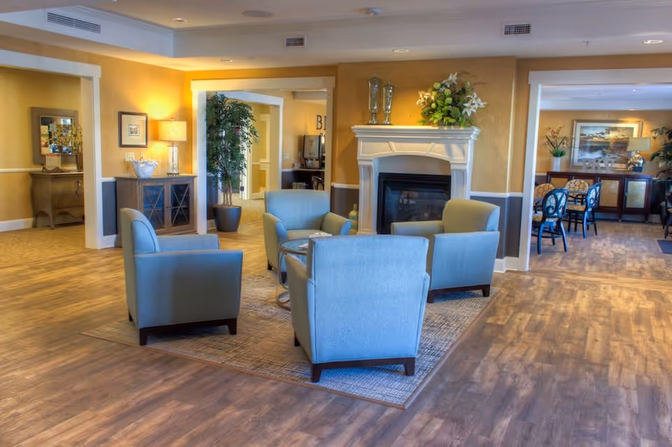 A cozy living room area in an assisted living facility featuring four light blue armchairs arranged around a small round glass table on a rug. The room has warm yellow walls with white trim, a white fireplace decorated with vases and flowers, and wood flooring. In the background, there is a dining area with tables and chairs, plants, and framed artwork on the walls.