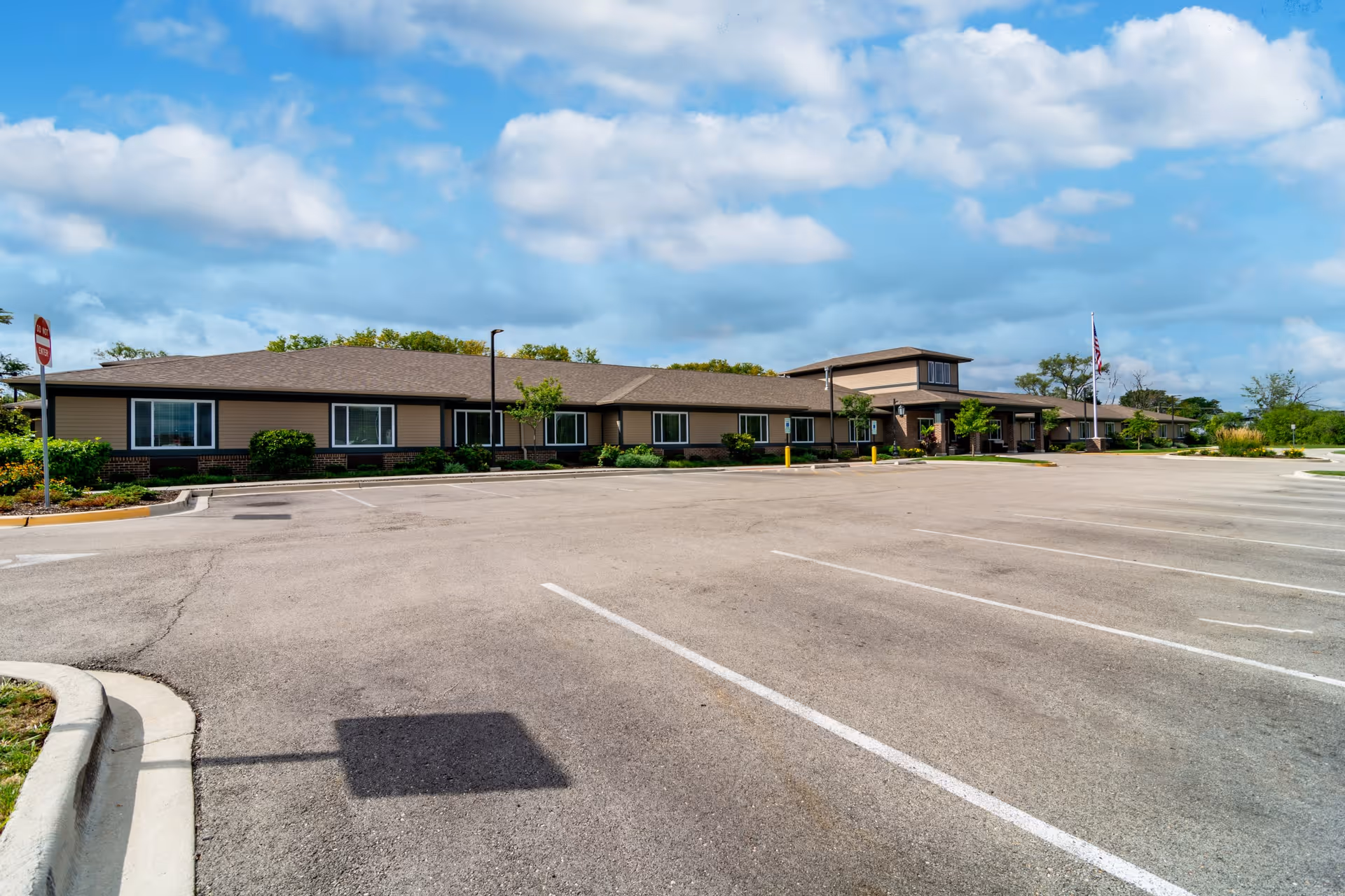 Wide view of the exterior of a single-story senior living facility building named Harvester Place with a large empty parking lot in front, some small trees and shrubs along the building, and a partly cloudy blue sky overhead.