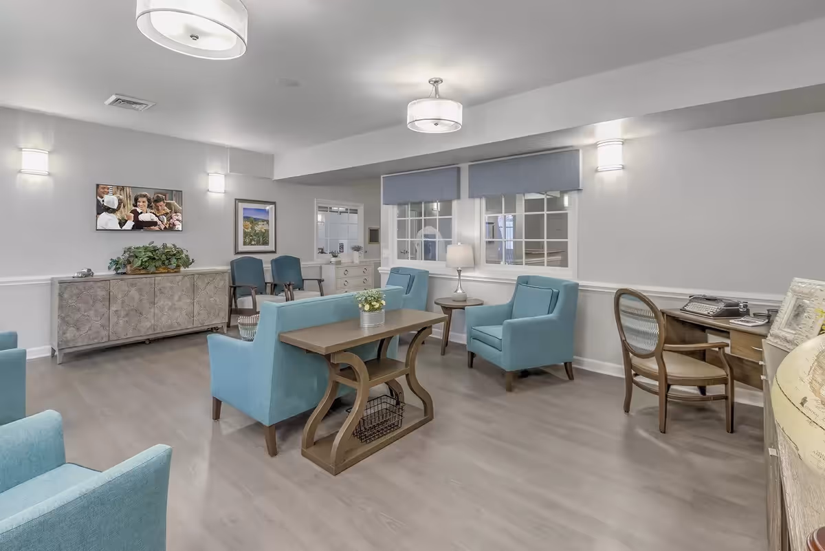 A bright and spacious living room area with light gray walls and wood flooring. The room features several light blue upholstered armchairs and a matching loveseat arranged around a wooden coffee table. There is a wooden side table with a lamp between two windows with blue valances. On the left wall, there is a decorative cabinet with a plant on top and a framed picture above it. Two additional chairs are placed near the cabinet. On the right side, there is a wooden desk with a chair and a vintage typewriter. The room is illuminated by ceiling lights and wall sconces.