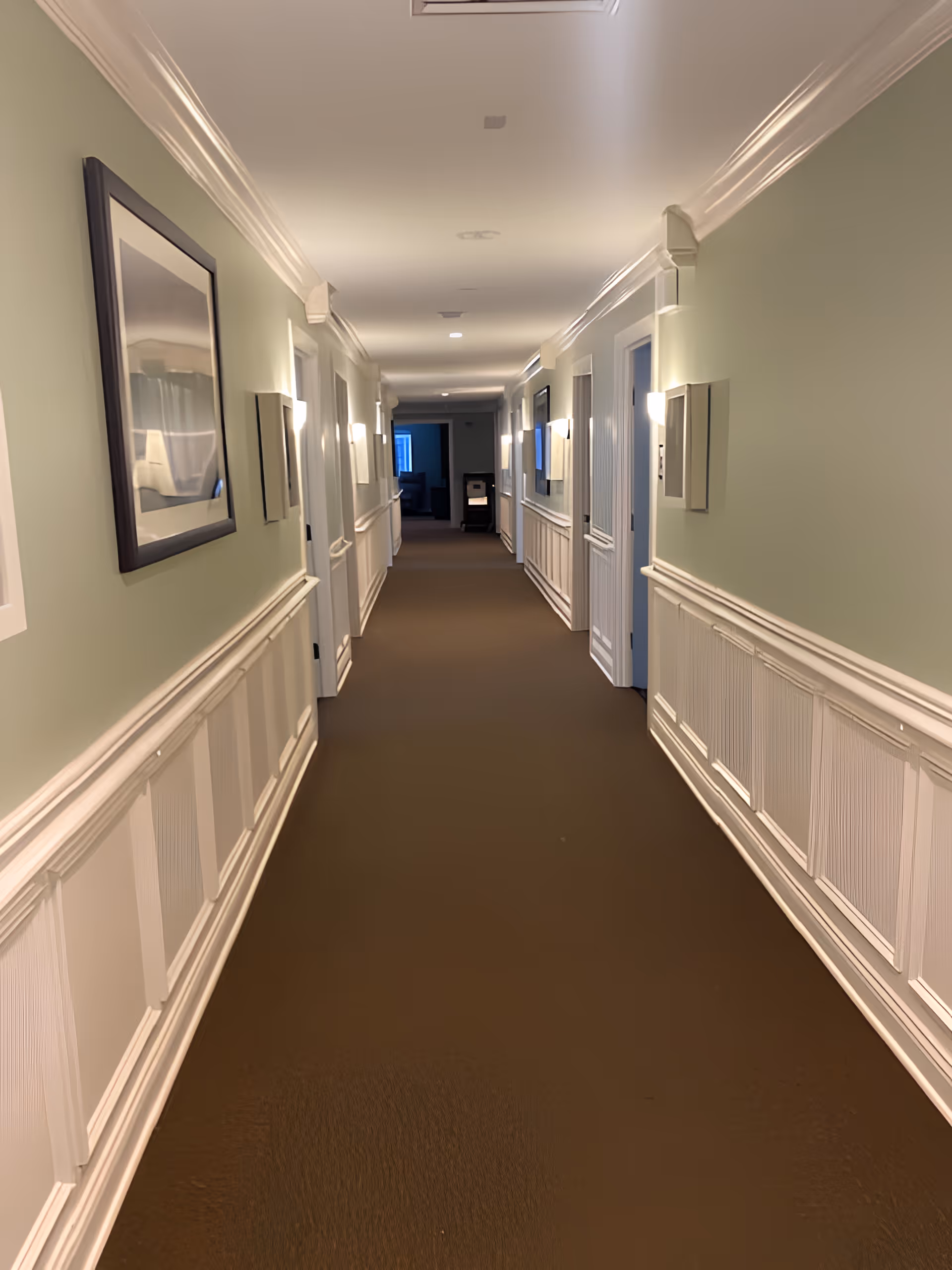 A long, well-lit hallway with light green walls and white wainscoting. Several closed white doors line both sides of the hallway, and framed artwork is hung on the walls. The floor is covered with a brown carpet, and wall sconces provide lighting along the corridor.