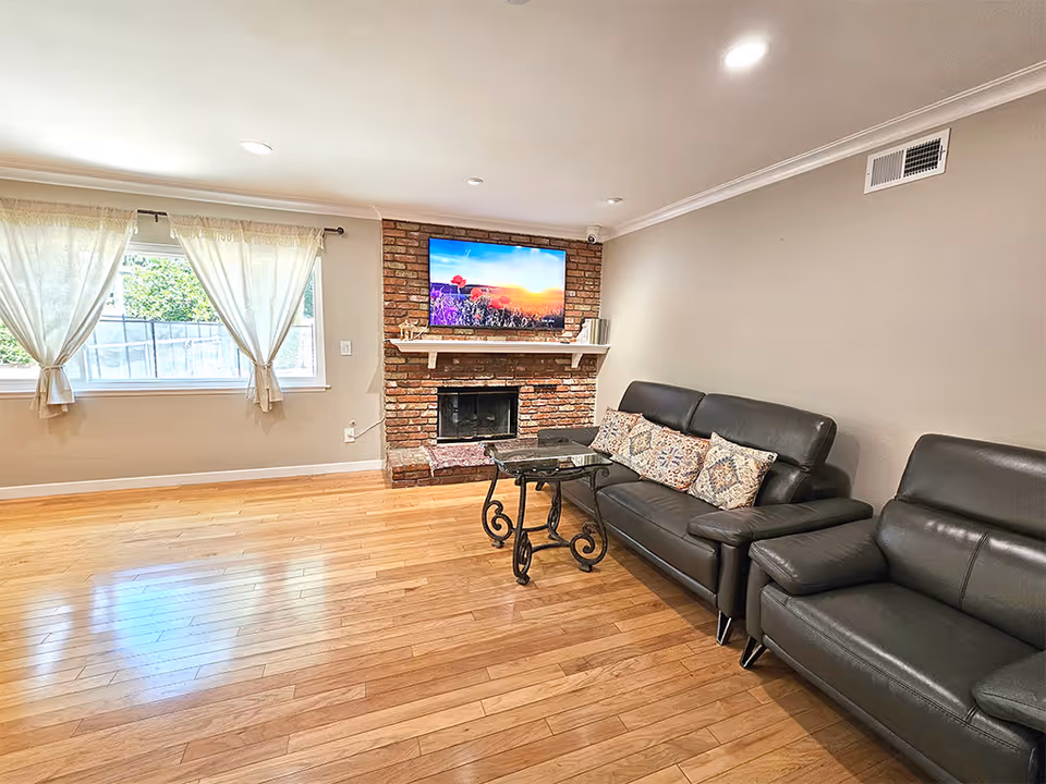 A bright living room with wooden flooring, a brick fireplace with a mounted flat-screen TV above it displaying a scenic image of a sunset and flowers. There are two black leather sofas with patterned cushions and a small glass coffee table with ornate metal legs. Light beige walls and a window with sheer white curtains letting in natural light.