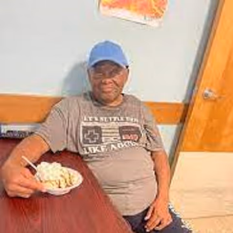 An elderly man wearing a blue cap and a gray t-shirt sits at a wooden table holding a bowl of food with a spoon. He is indoors, with a light blue wall, a wooden door, and a colorful drawing or poster on the wall behind him.
