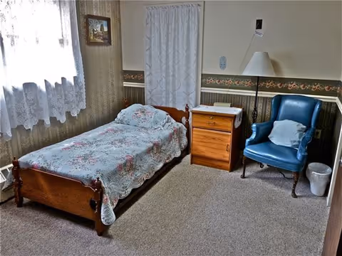 A small bedroom with a single wooden bed covered with a floral quilt, a wooden nightstand with a lamp and a doily, a blue upholstered armchair with a pillow, a trash bin, and a window with lace curtains letting in natural light.