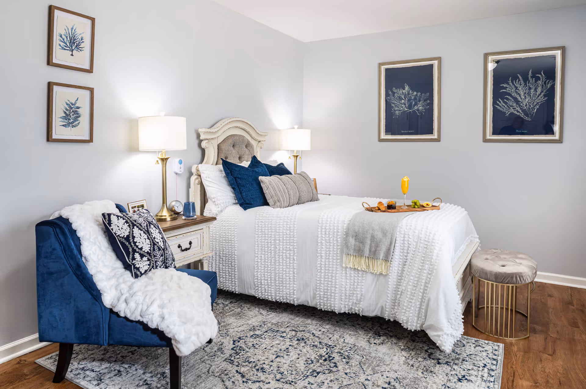 Staged bedroom with a white textured bed topped with blue pillows, a navy accent chair, bedside lamps, framed wall art, and a breakfast tray on the bed.