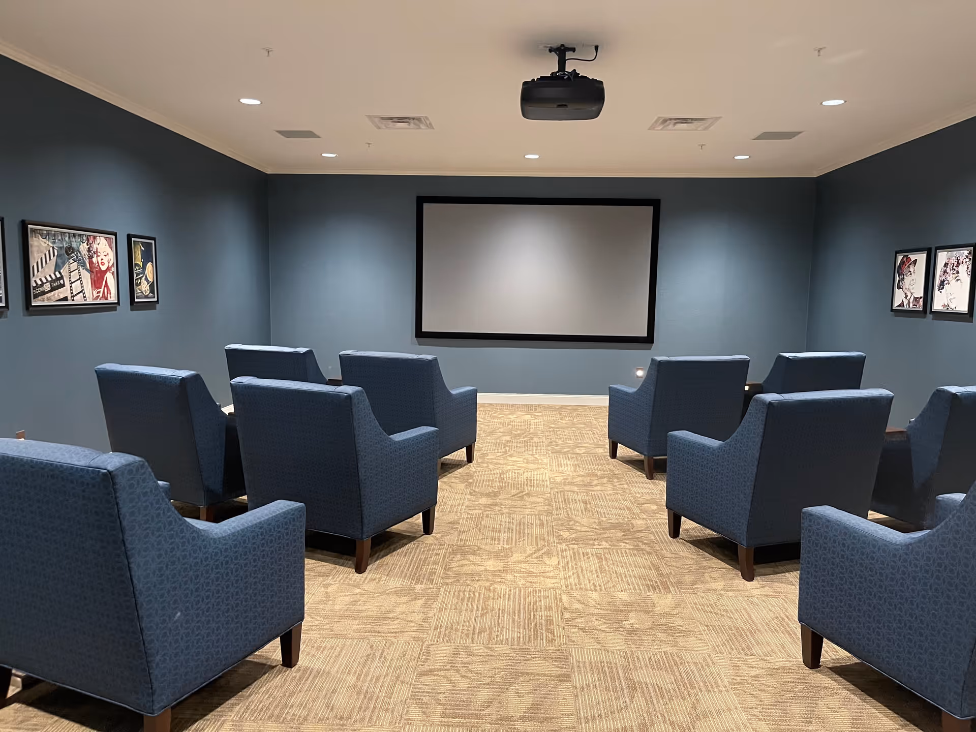 A small theater room with eight blue upholstered armchairs arranged in two rows facing a large blank projector screen on a blue wall. The room has beige carpet flooring and framed artwork on the side walls. A ceiling-mounted projector is visible above the chairs.