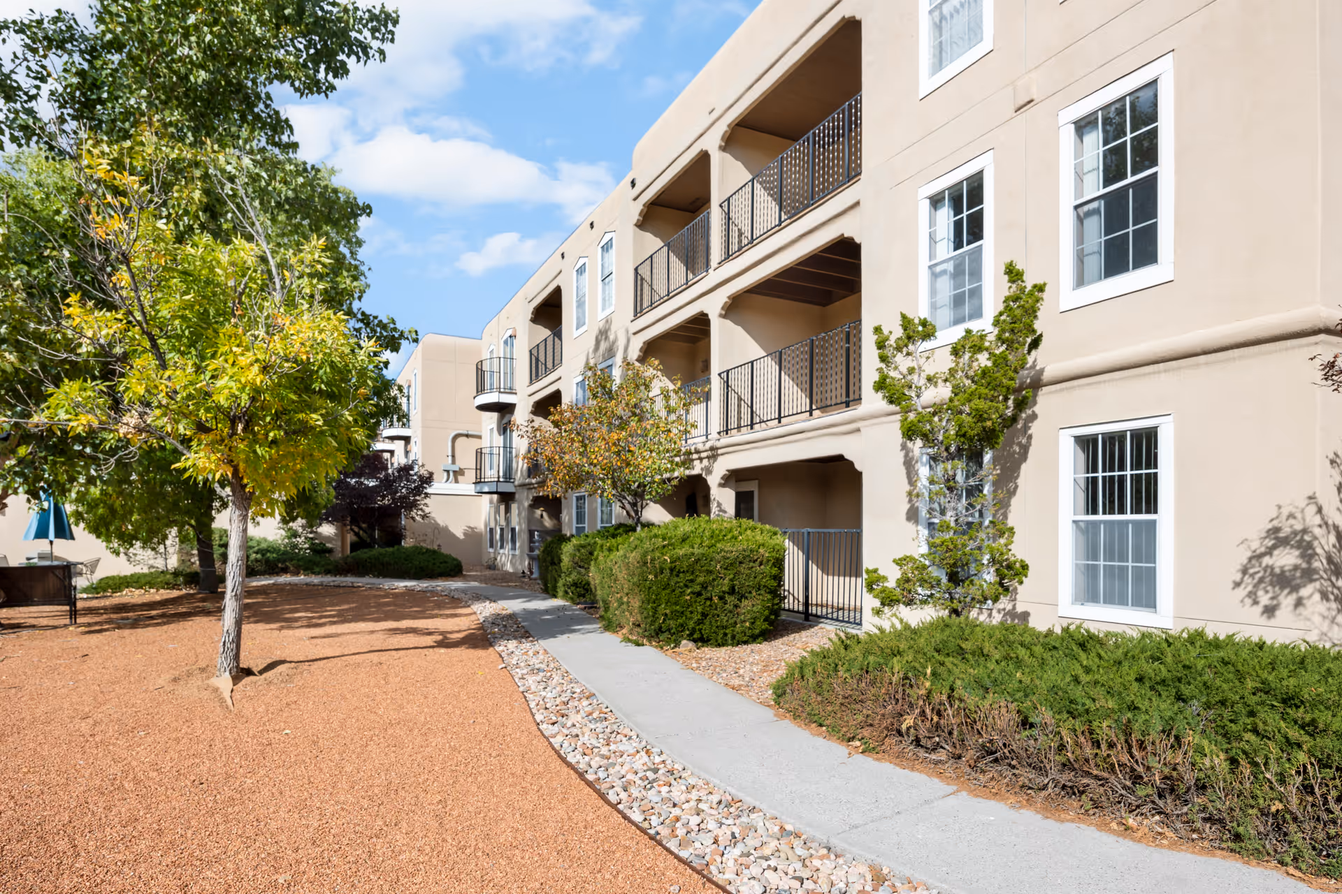Exterior view of a beige multi-story residential building with balconies and large windows, surrounded by landscaped grounds with trees, bushes, and a gravel pathway under a partly cloudy sky.