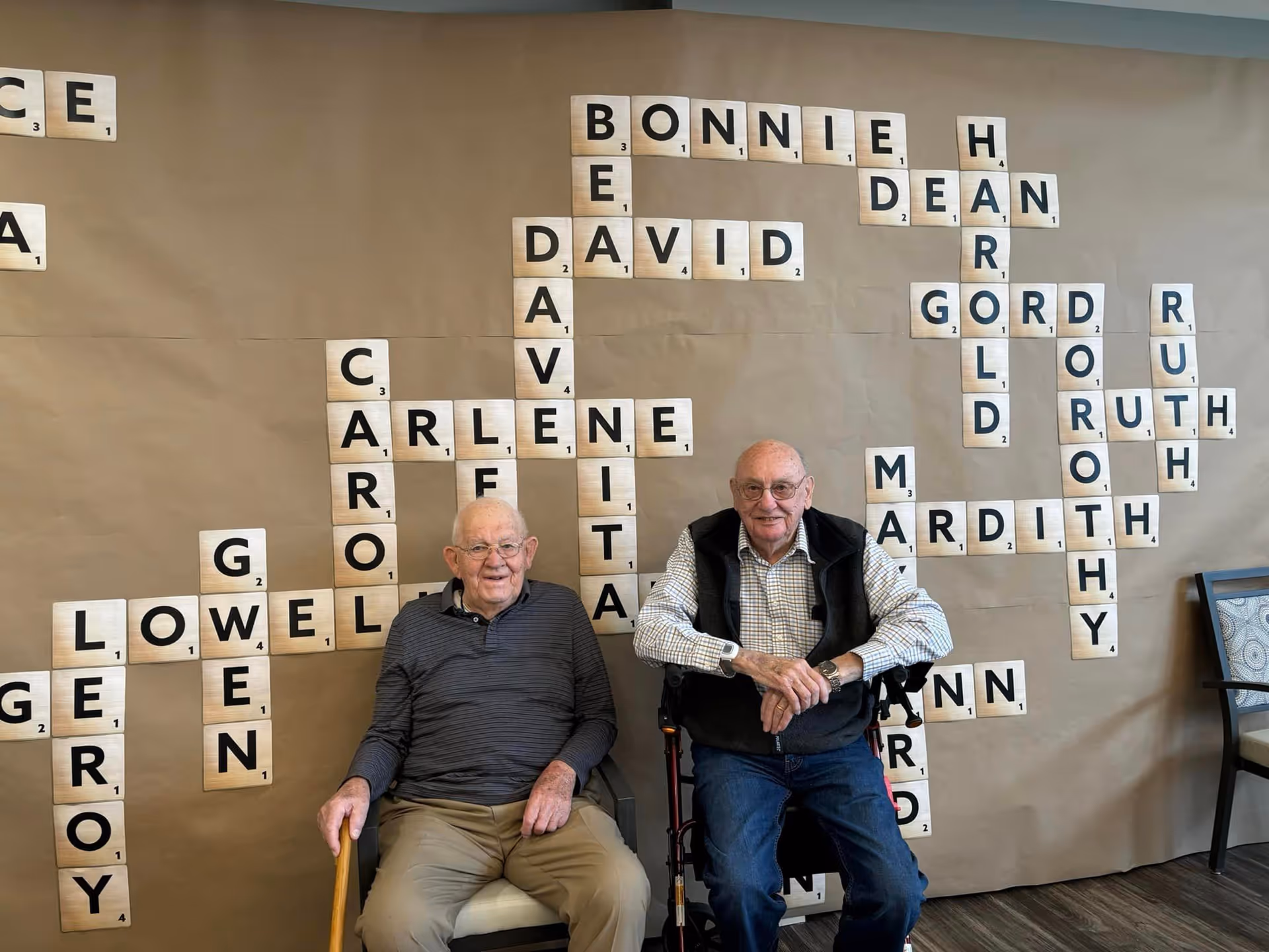 Two elderly men sitting in chairs in front of a wall decorated with large Scrabble-style letter tiles forming names in a crossword pattern. One man is holding a cane and the other has a walker. There is a chair to the right side of the image.
