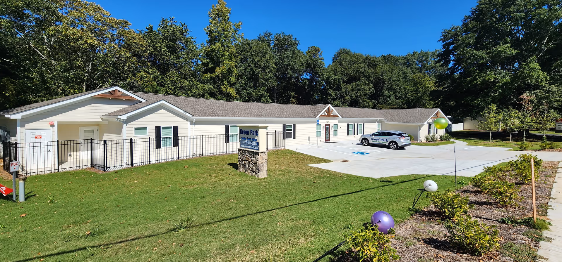 Exterior view of Green Park Senior Care Home, a single-story building with light-colored siding and a gray roof. There is a parking lot with a car displaying the facility's name, a green lawn, some bushes, and trees in the background under a clear blue sky.