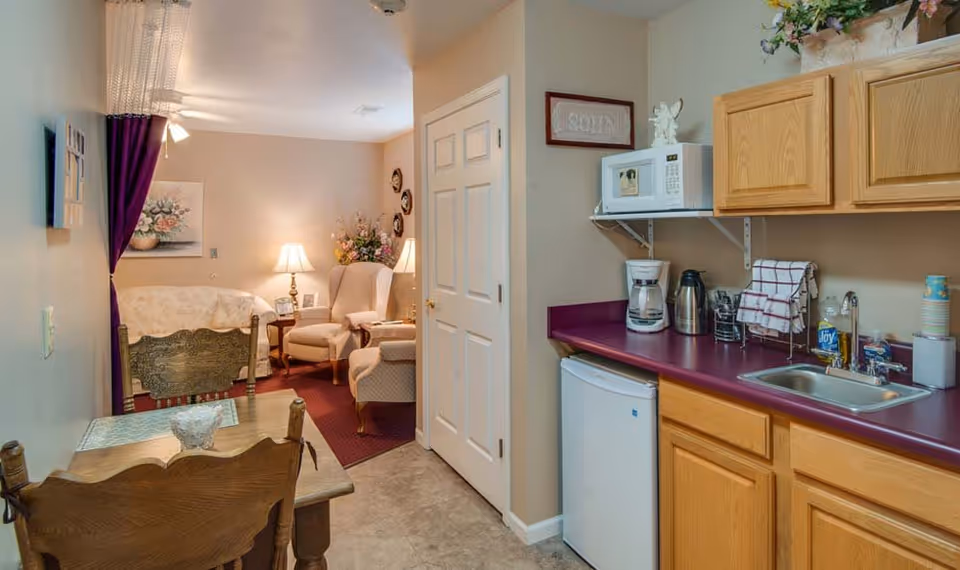 Small kitchenette with wooden cabinets, microwave, sink and mini-fridge opening to a cozy sitting area with armchairs and a lamp.