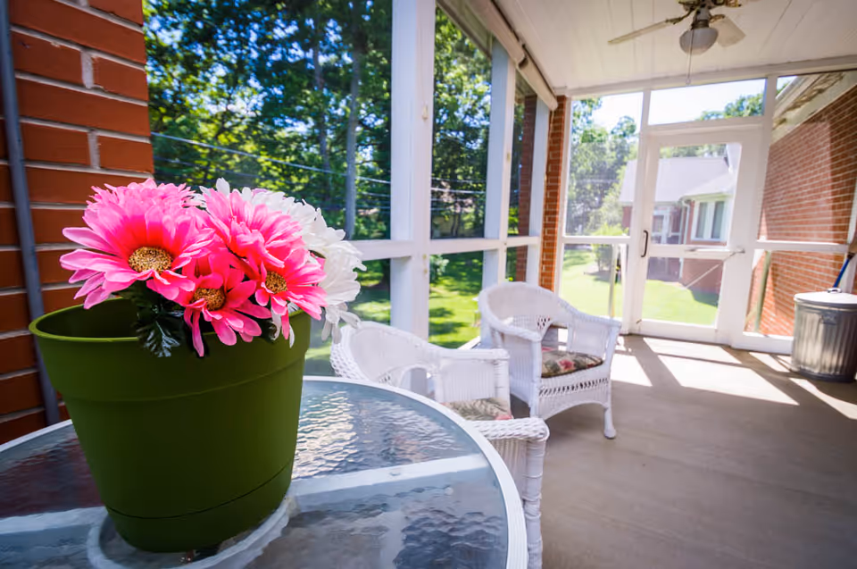 A bright screened-in porch with two white wicker chairs with floral cushions and a glass table holding a green pot with pink and white flowers. The porch overlooks a green lawn and trees outside.