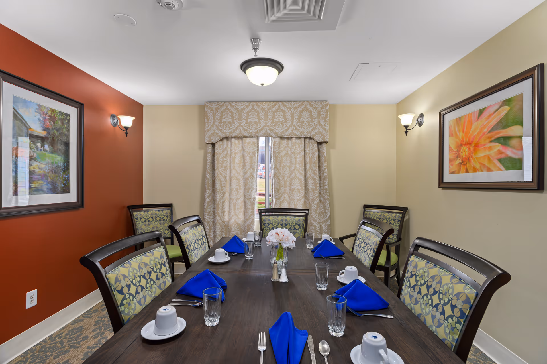Dining room with a long table set with blue napkins, cups and glasses, surrounded by upholstered chairs and framed artwork.