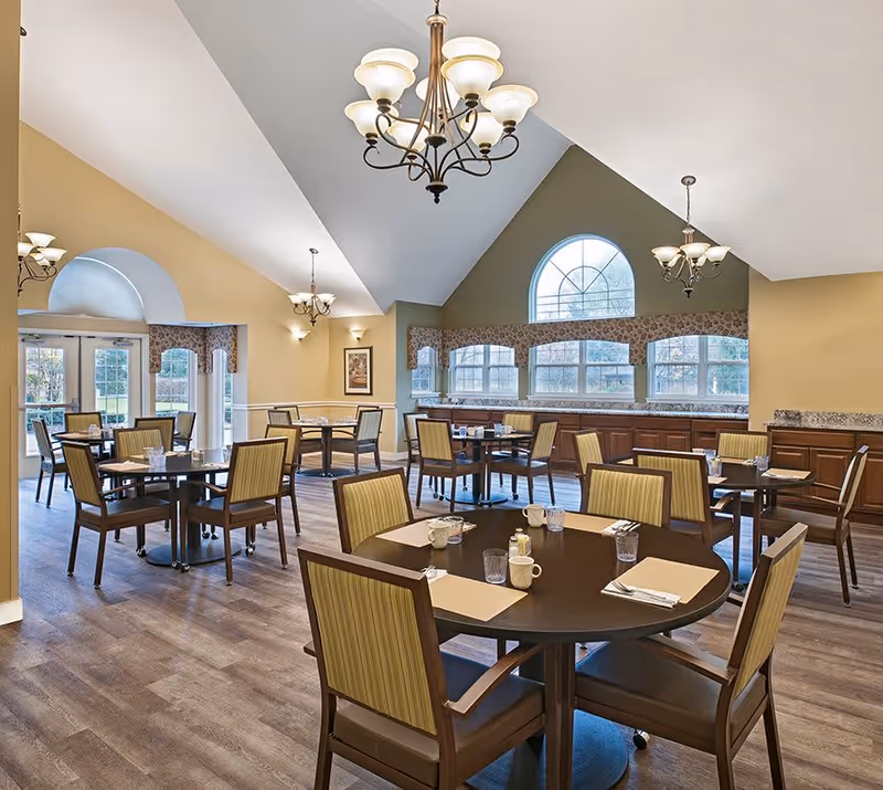 A spacious dining room in a senior living facility with multiple round tables set with placemats, cups, glasses, and utensils. The room features high vaulted ceilings with elegant chandeliers, large windows with decorative valances, and wooden flooring. The walls are painted in warm tones, and there is ample natural light coming through the windows.