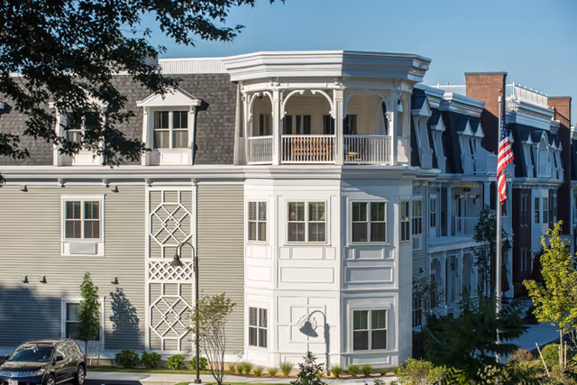Exterior view of a multi-story senior living facility building with white and gray siding, multiple windows, a covered balcony, and an American flag on a flagpole in front. There are trees, shrubs, and parked cars visible around the building under a clear blue sky.