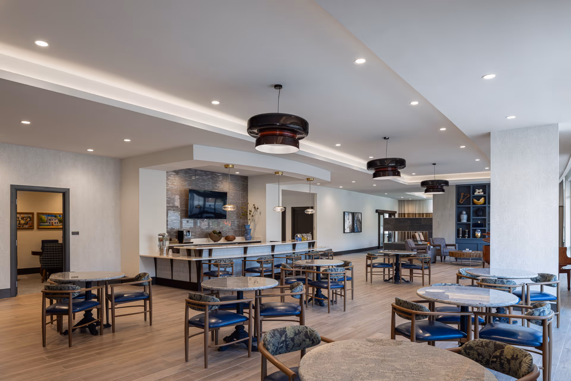 A spacious and modern dining area in a senior living facility with multiple round tables and chairs arranged neatly. The room features a bar counter with stools, a mounted TV, pendant lights, and decorative shelving. The flooring is wooden, and the ceiling has recessed lighting and stylish hanging fixtures.