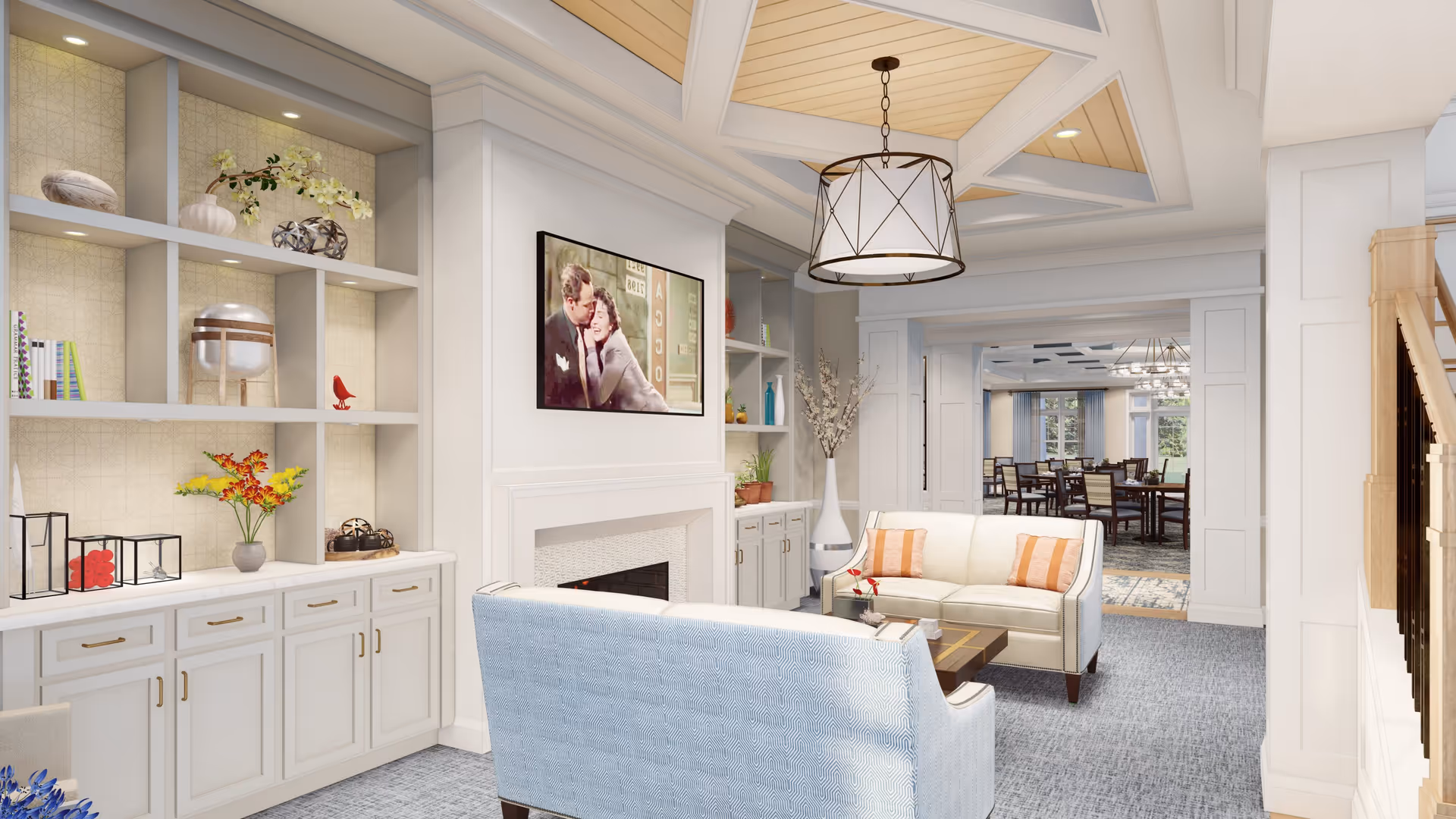 A cozy senior living facility common area with a light blue patterned sofa and a cream-colored sofa with orange striped pillows facing a fireplace with a mounted TV above it. Built-in shelves with decorative items and plants flank the fireplace. The ceiling features a wooden panel design with a modern chandelier. In the background, a dining area with tables and chairs is visible through a wide doorway.