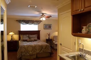 A cozy bedroom with a bed covered in patterned bedding, a nightstand with a lamp, a window with a valance, a ceiling fan with lights, and a small kitchenette area with a sink and cabinets.
