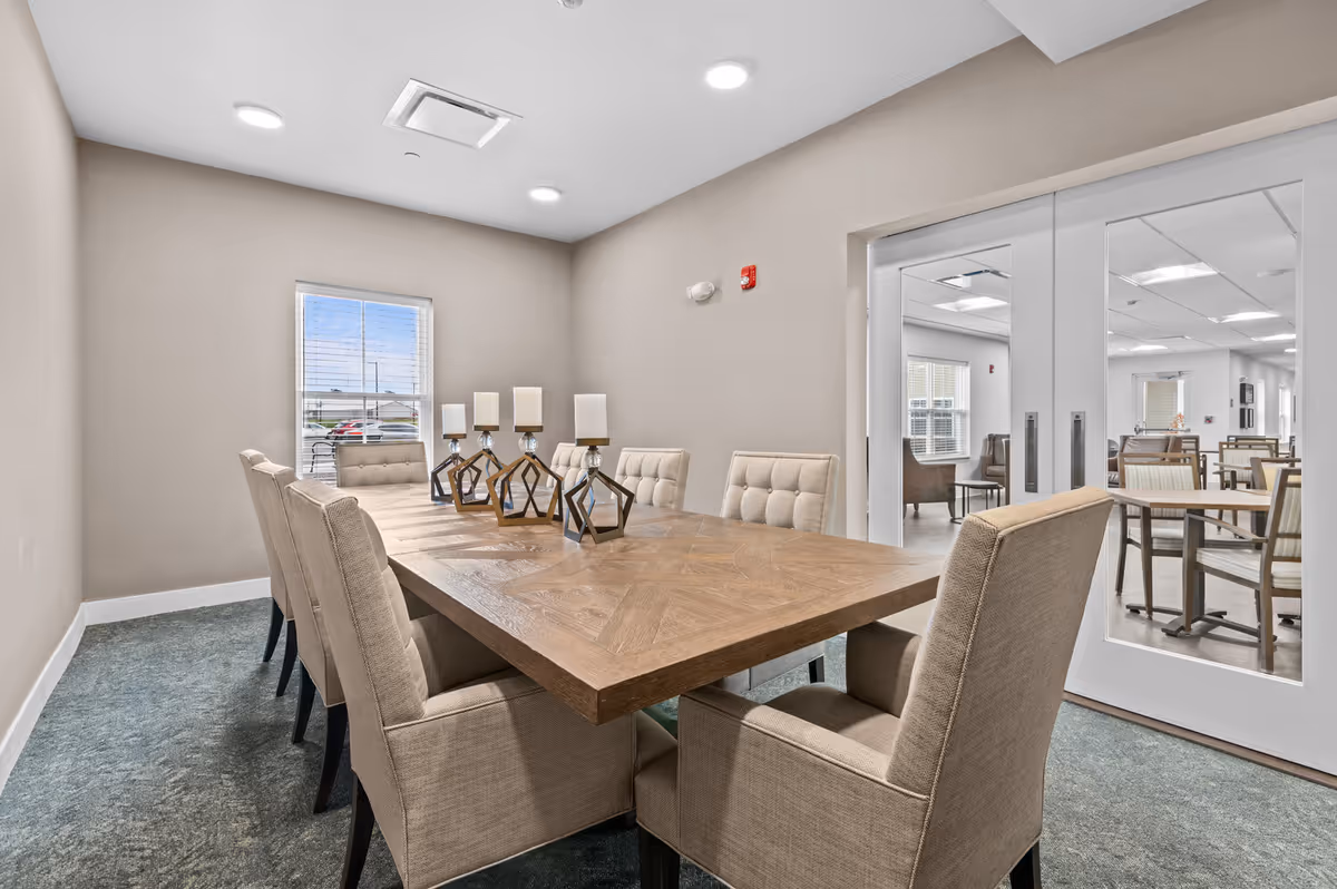 A modern dining room with a large wooden table surrounded by eight beige upholstered chairs. The table is decorated with five geometric candle holders. There is a window on the far wall letting in natural light, and double glass doors open to another room with additional tables and chairs.