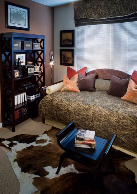 Cozy living room area with a patterned daybed adorned with multiple pillows in shades of brown and red, a dark wooden bookshelf filled with books and decorative items, a small black coffee table with a stack of books on it, and a cowhide rug on the floor. The window behind the daybed has blinds and a decorative valance.