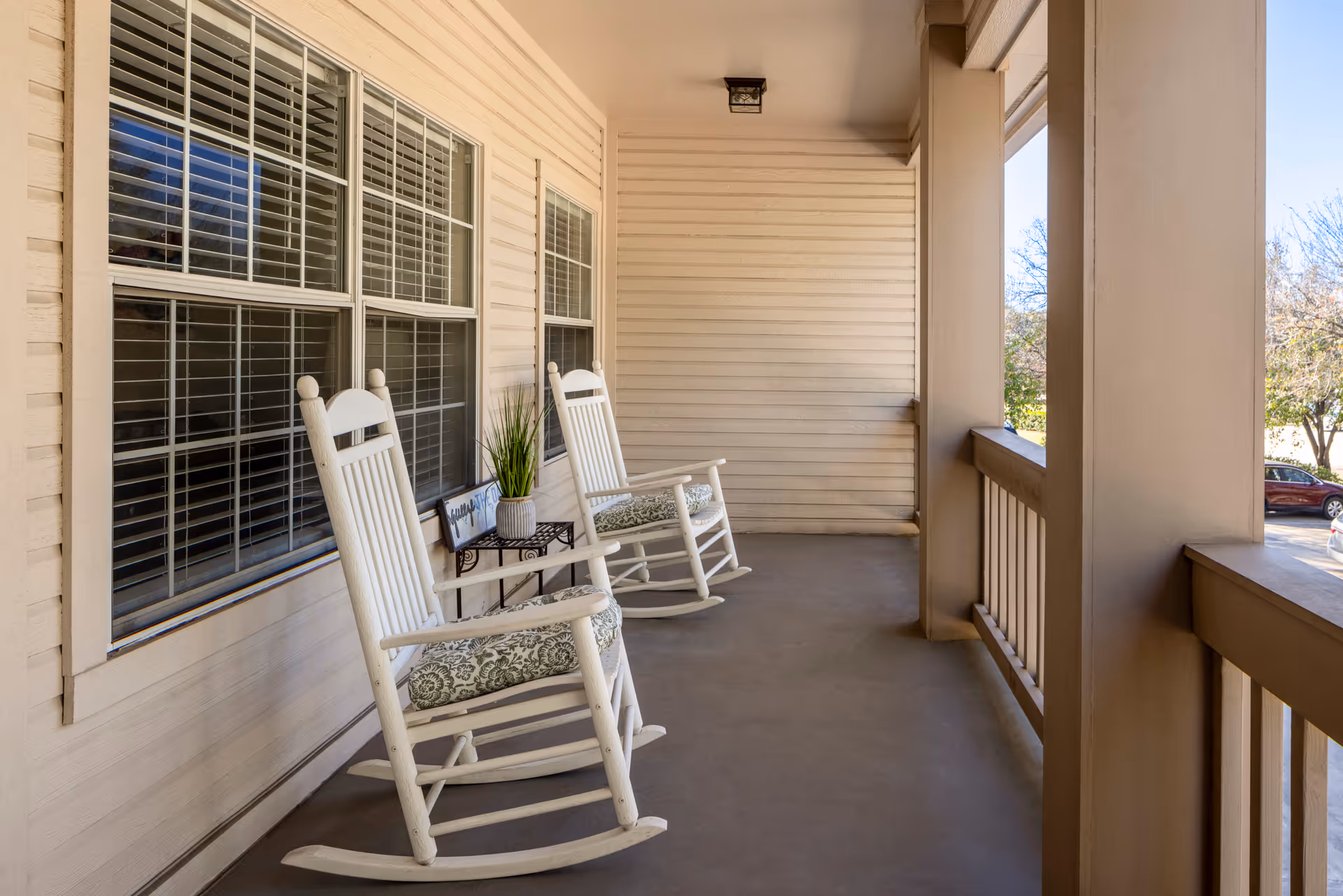 A covered porch area with two white wooden rocking chairs with patterned cushions, a small black metal side table with a potted plant and a decorative sign, beige siding walls, and large windows with white shutters. The porch overlooks a parking area with some trees and cars visible in the background.