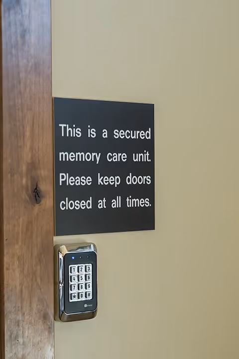 A close-up view of a door with a keypad lock and a black sign that reads 'This is a secured memory care unit. Please keep doors closed at all times.'