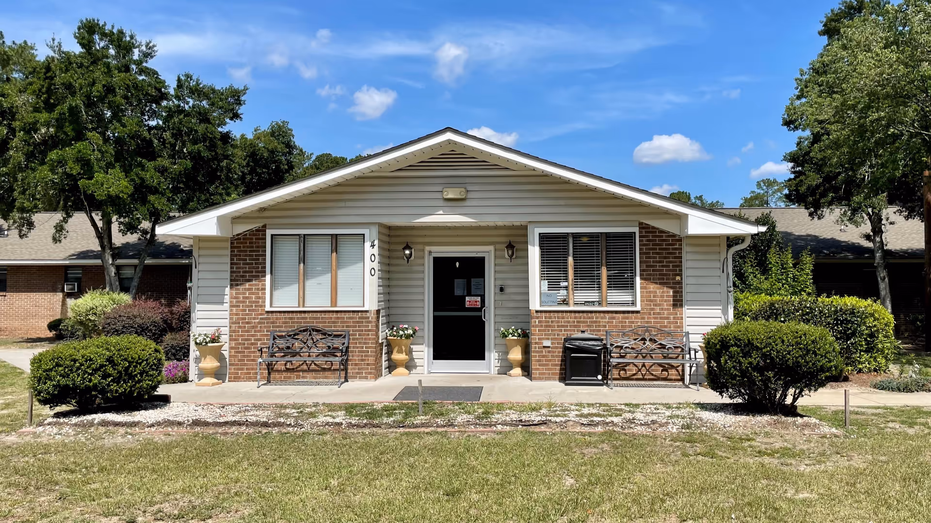 Front exterior view of a single-story nursing and rehabilitation center building with brick and white siding, two benches on the porch, potted plants, and bushes on either side under a blue sky with some clouds.