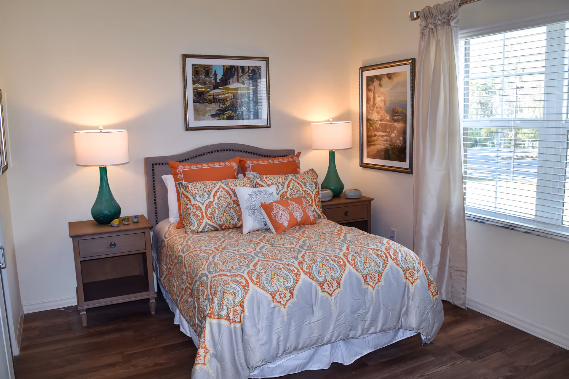 A cozy bedroom with a bed dressed in an orange and white patterned comforter and multiple pillows. There are two wooden nightstands on either side of the bed, each with a green lamp. The walls are light-colored and decorated with framed artwork. A window with white blinds and light-colored curtains lets in natural light.