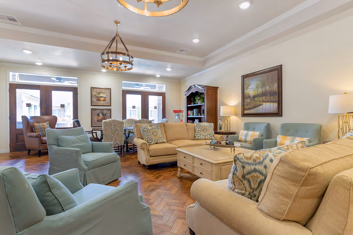 A bright and spacious senior living facility common area featuring comfortable beige and light blue upholstered sofas and armchairs arranged around a wooden coffee table. The room has wooden parquet flooring, a wooden cabinet with decorative items, two large windows with wooden frames, and warm lighting from ceiling fixtures and table lamps. A framed landscape painting hangs on the wall.