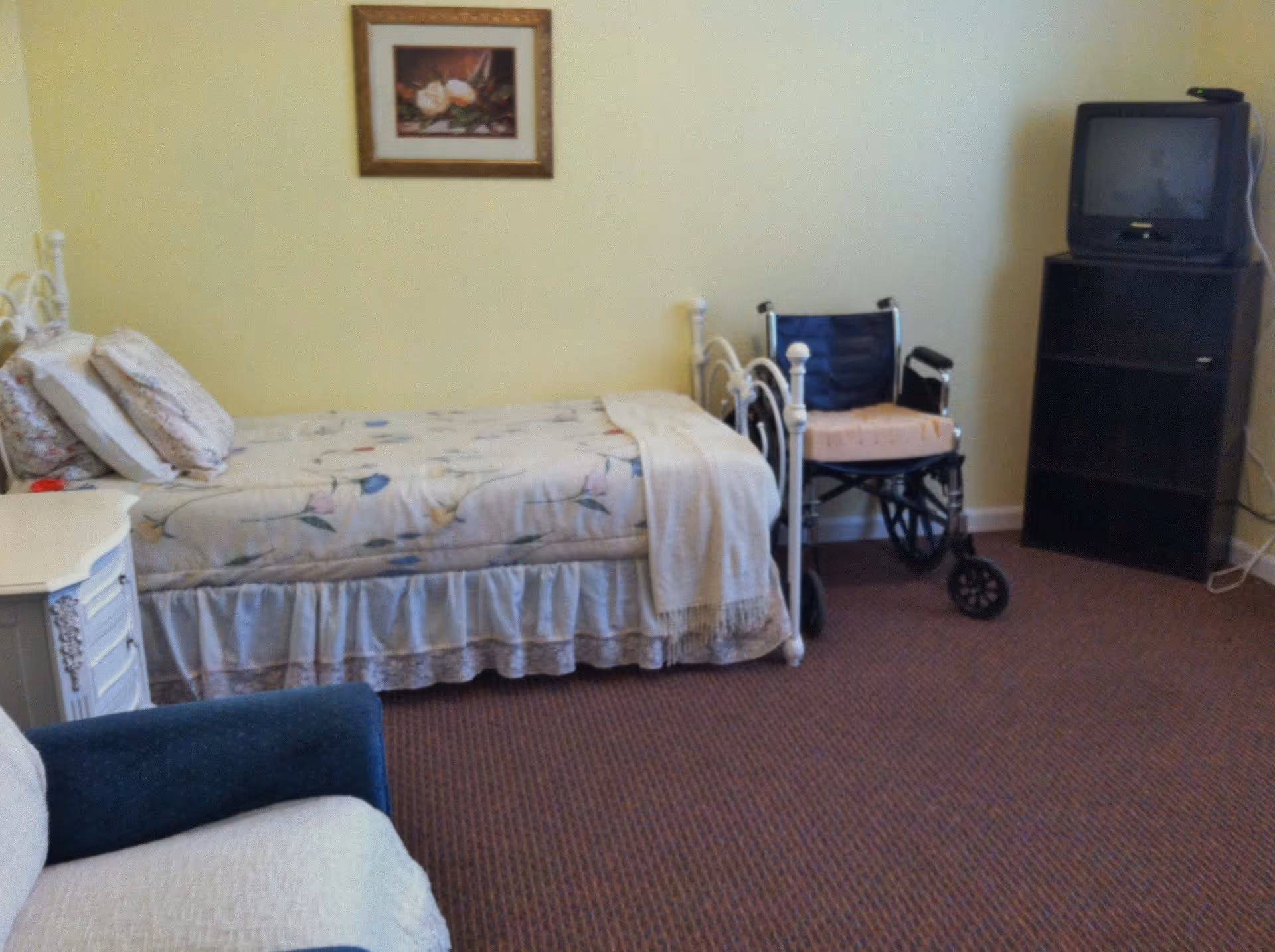 A small bedroom with a single bed covered with floral bedding and pillows, a wheelchair next to the bed, a dark wooden shelving unit with an old-style TV on top, and a blue armchair partially visible in the foreground. The walls are painted light yellow and there is a framed floral picture hanging above the bed.