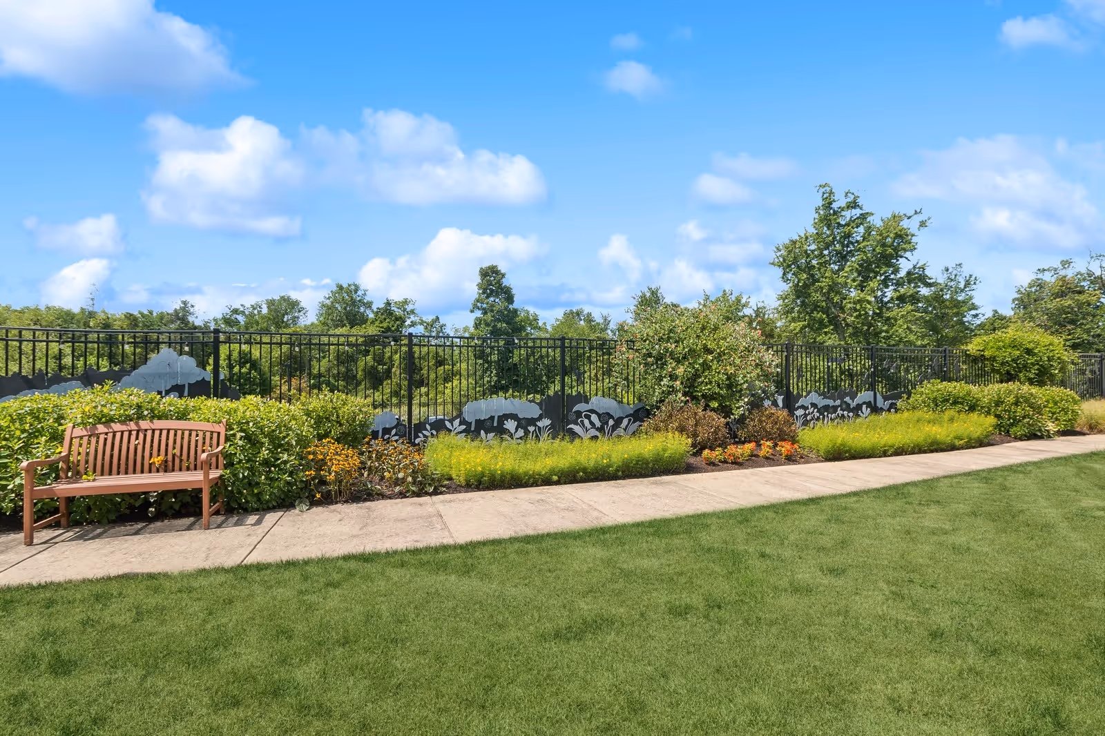 A peaceful outdoor garden area with a wooden bench on a concrete pathway, surrounded by green bushes, colorful flowers, and a decorative black metal fence with floral patterns. The sky is blue with scattered white clouds.