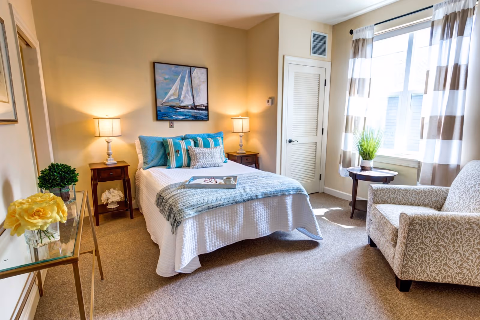 Sunlit furnished bedroom with a neatly made bed flanked by bedside tables and lamps, an armchair by a window, and coastal artwork on the wall.
