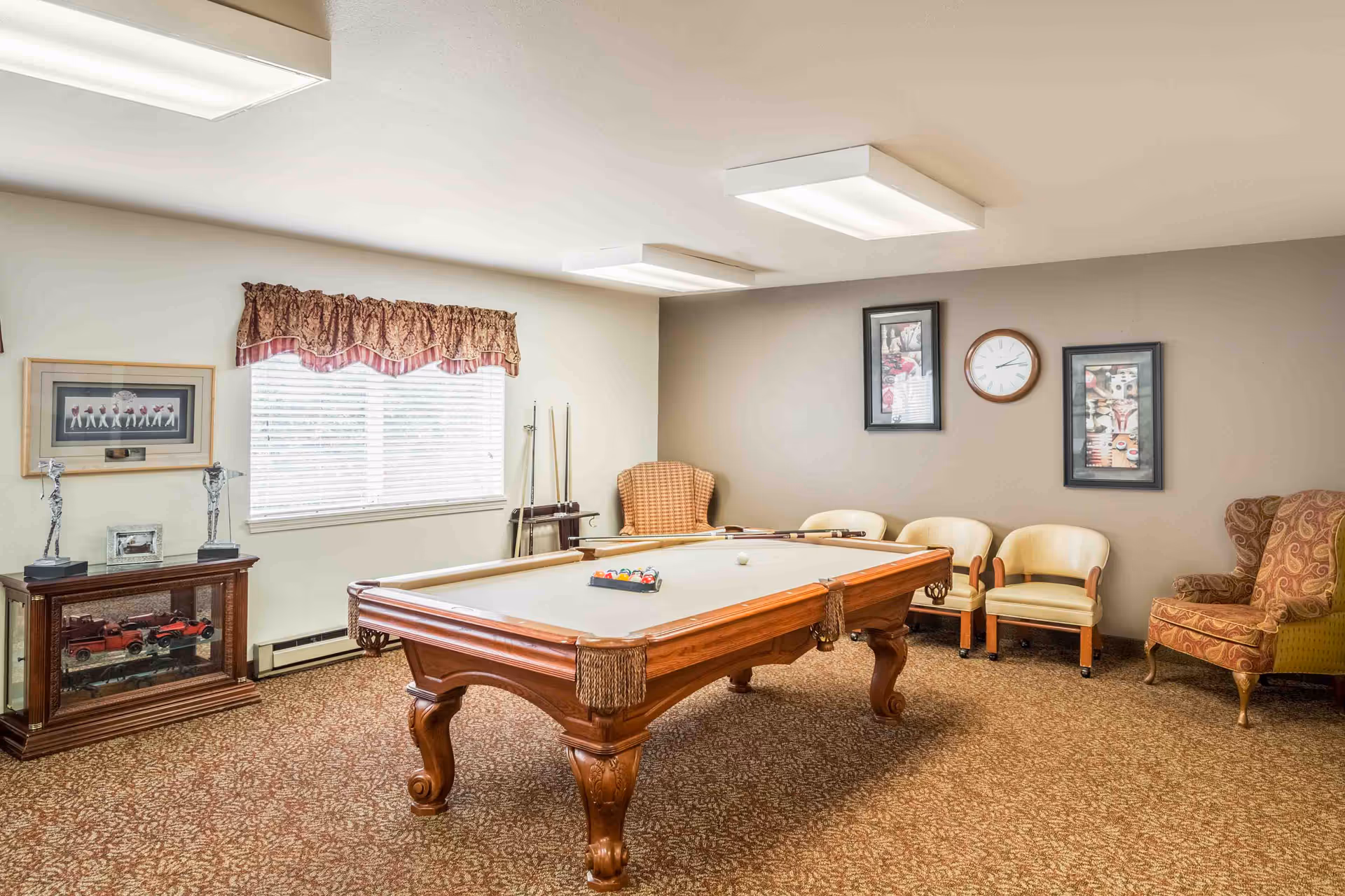 A recreational room with a wooden pool table in the center, a rack with pool cues against the wall, several chairs including two upholstered armchairs and three cream-colored chairs, a window with blinds and a valance, framed pictures and a clock on the wall, and a cabinet displaying model cars and trophies.