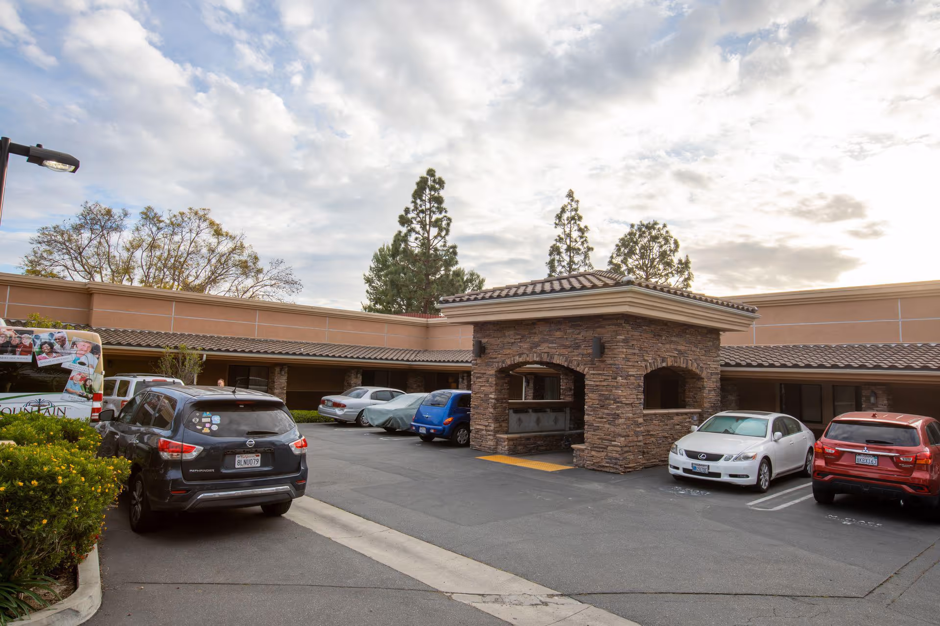 Parking lot area of a senior living facility with several parked cars and a stone-covered entrance structure under a partly cloudy sky.