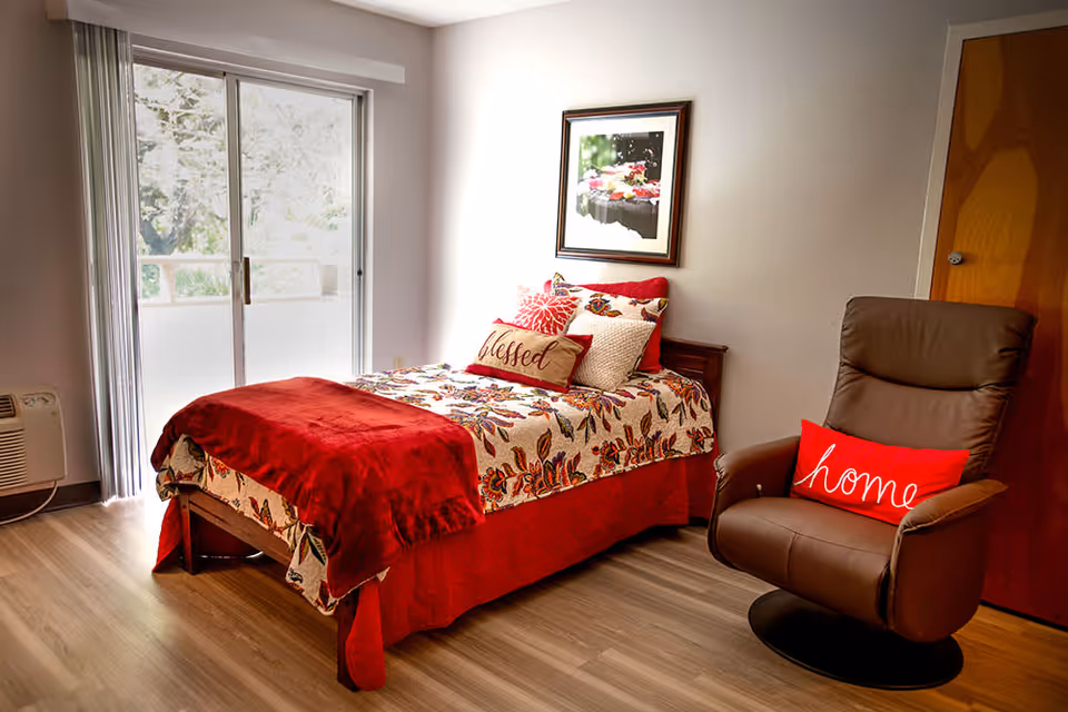 A cozy bedroom with a single bed covered in a floral patterned bedspread and a red throw blanket. The bed has decorative pillows, including one with the word 'blessed'. Next to the bed is a brown leather recliner chair with a red pillow that says 'home'. There is a wooden door on the right side and a large sliding glass door on the left letting in natural light. A framed picture hangs on the wall above the bed.