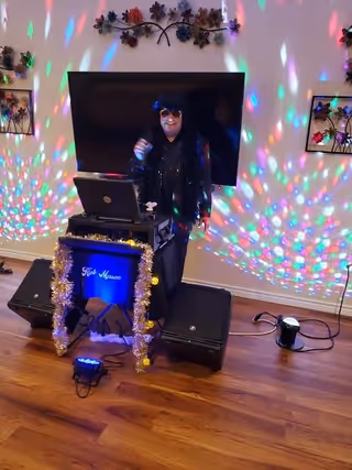 A person dressed in black with sunglasses stands behind a DJ setup in a room with wooden floors and a large flat-screen TV mounted on the wall. Colorful disco lights create patterns on the walls and floor. Decorative metal wall art with flowers is visible above and beside the TV.