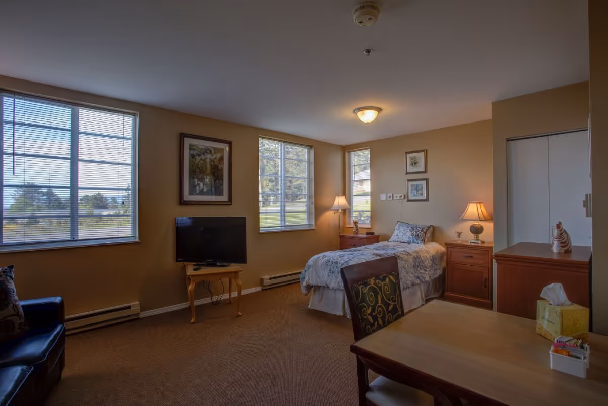 Well-lit assisted living bedroom with a single bed, nightstands, a TV, windows, and a small table and chair.