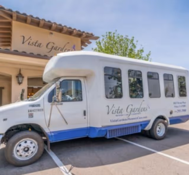 A white and blue shuttle bus parked in front of a building with a sign that reads 'Vista Gardens'. The bus has the Vista Gardens logo and website on its side. The building has a tiled roof and a lantern-style light fixture near the entrance.