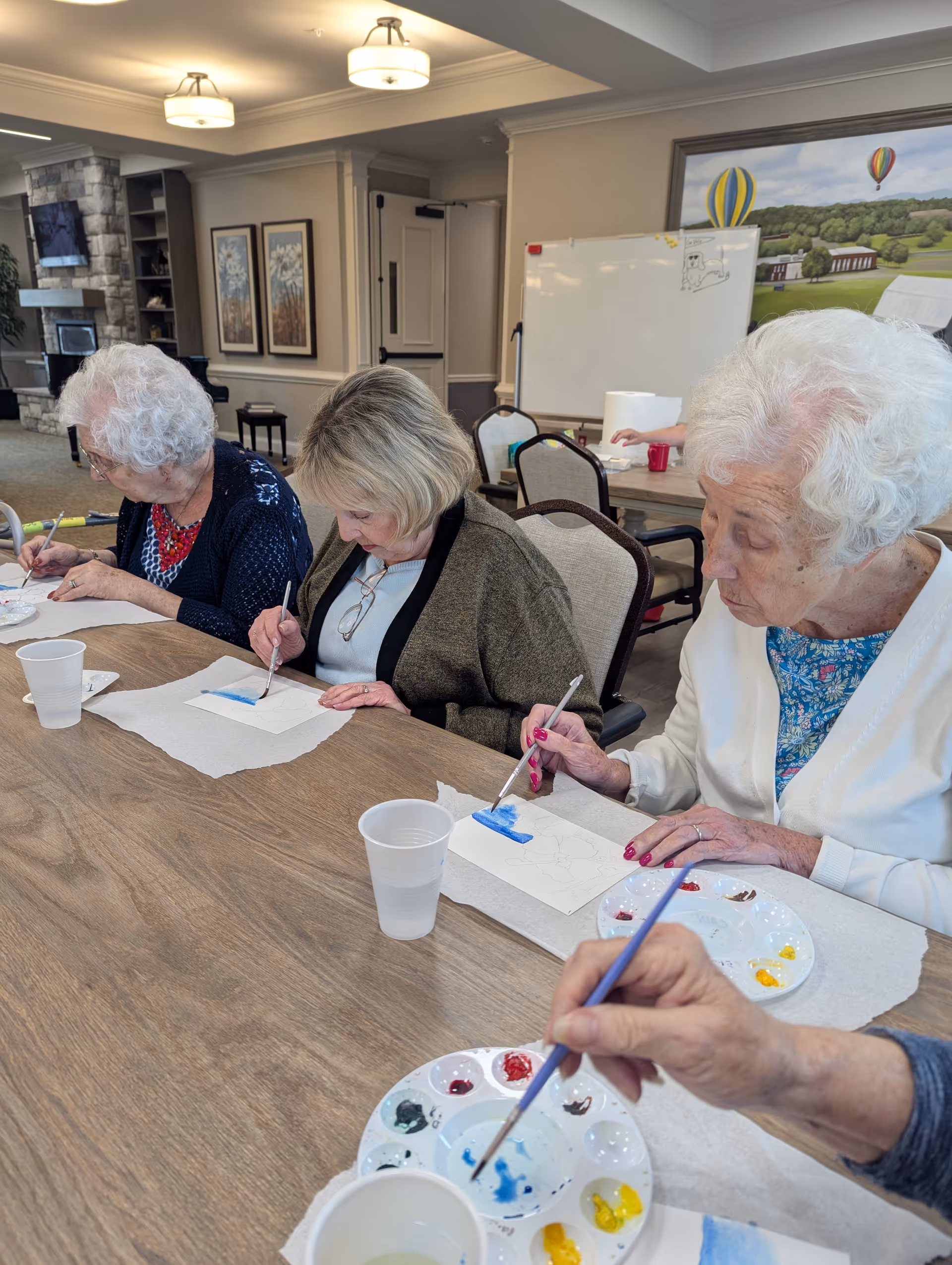 Four elderly women seated at a wooden table in a well-lit room, engaged in painting on small sheets of paper with paintbrushes and palettes of various colors. The room has framed artwork on the walls, a whiteboard, and a large painting of hot air balloons in the background.