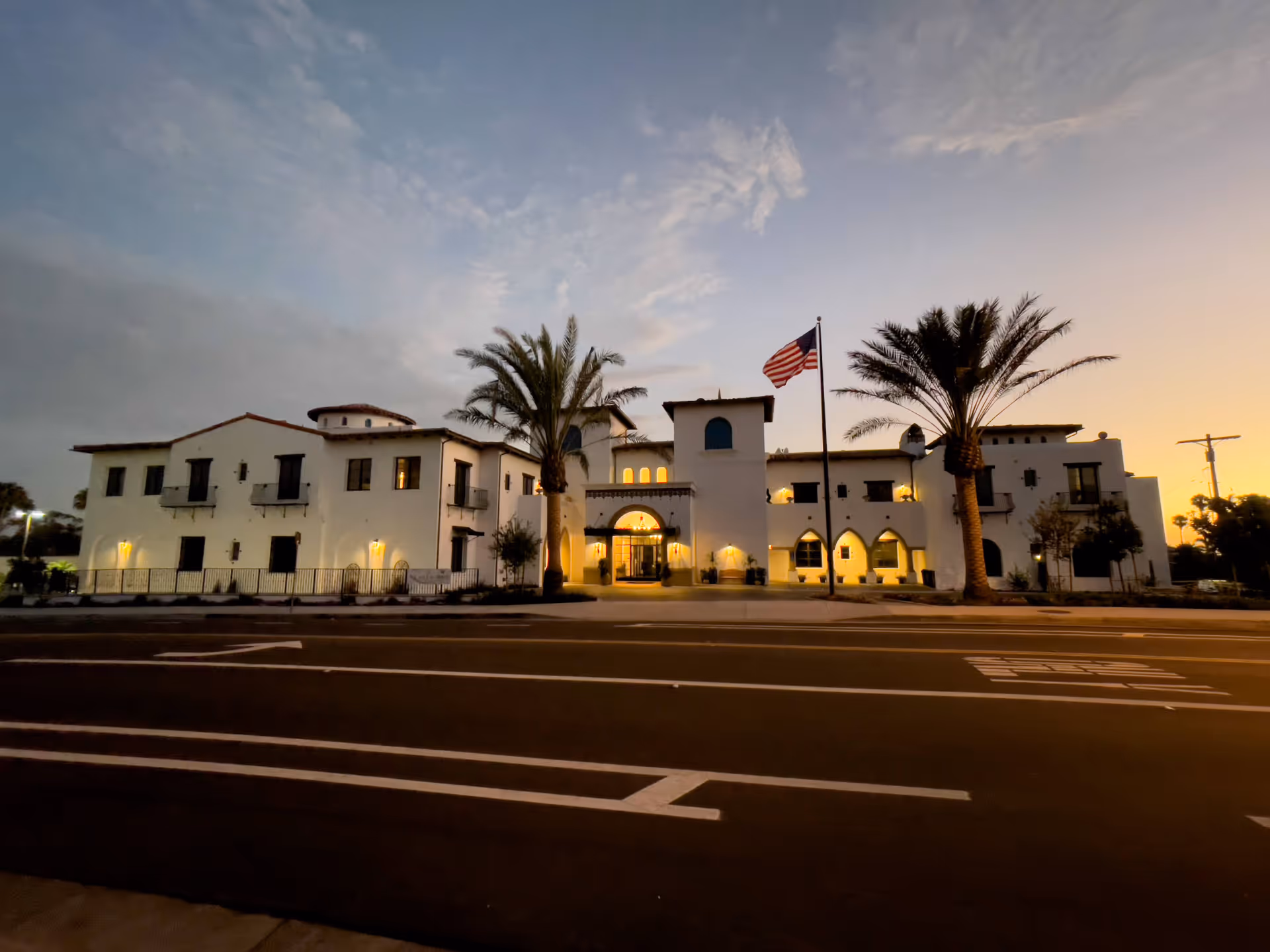 Exterior front view of a large, white, two-story building with Spanish-style architecture at dusk. The building is illuminated with warm lights and features palm trees and an American flag in front. The sky is partly cloudy with a sunset glow.