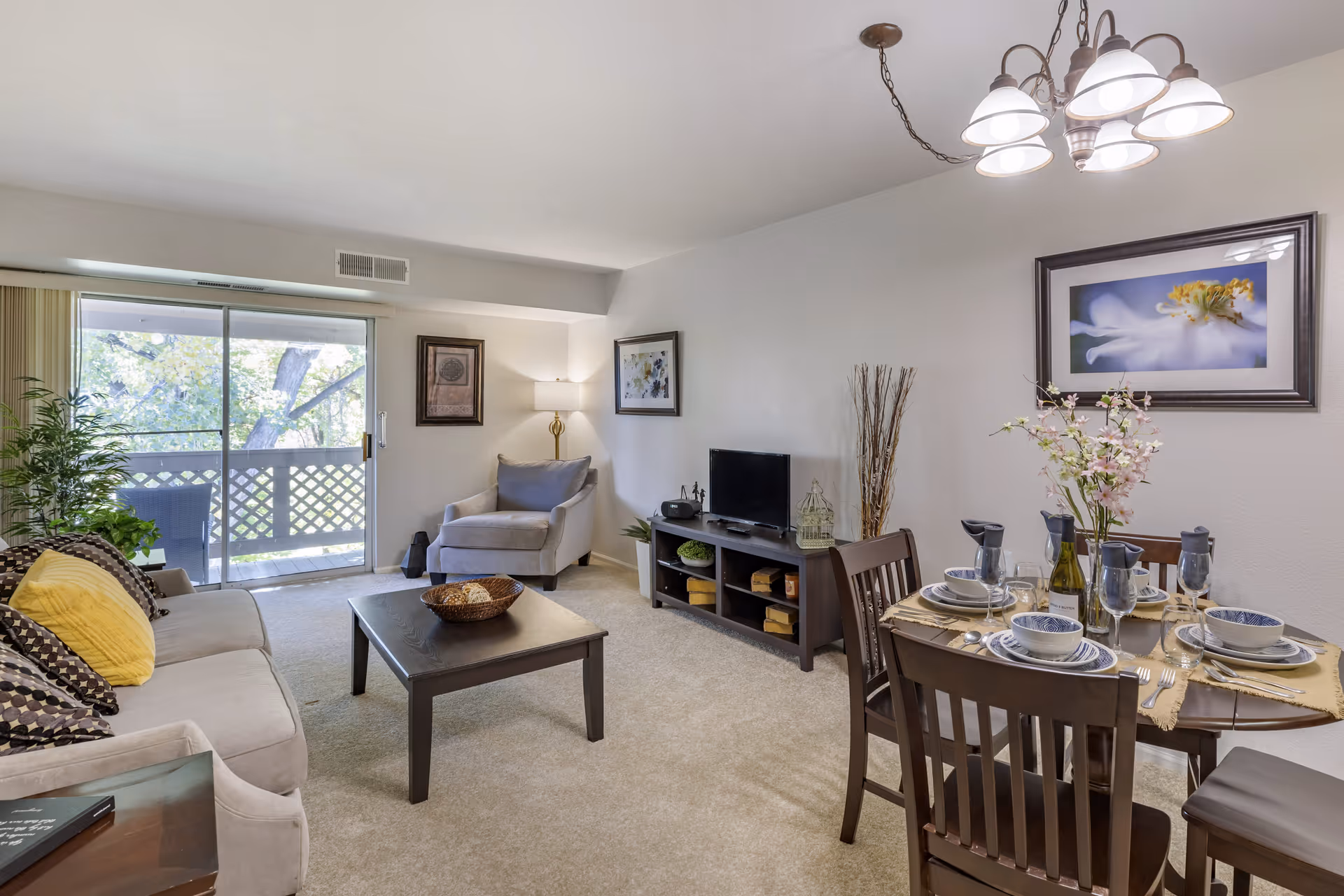 A bright and cozy living room and dining area with a beige sofa adorned with patterned and yellow pillows, a wooden coffee table with a decorative bowl, a gray armchair, a TV on a dark wooden stand, and a dining table set for four with plates, bowls, glasses, and a floral centerpiece. The room has carpeted floors, framed artwork on the walls, a floor lamp, and a sliding glass door leading to a balcony with outdoor seating and greenery visible outside.