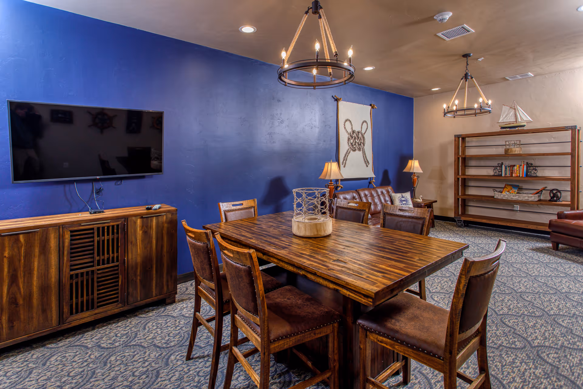 A cozy common area with a wooden table surrounded by six chairs, a blue accent wall with nautical-themed decor, a flat-screen TV mounted above a wooden cabinet, two chandeliers hanging from the ceiling, a brown leather couch with pillows, and a wooden bookshelf with decorative items.