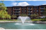 Brick senior living campus building with balconies, green trees, and a central fountain pond.