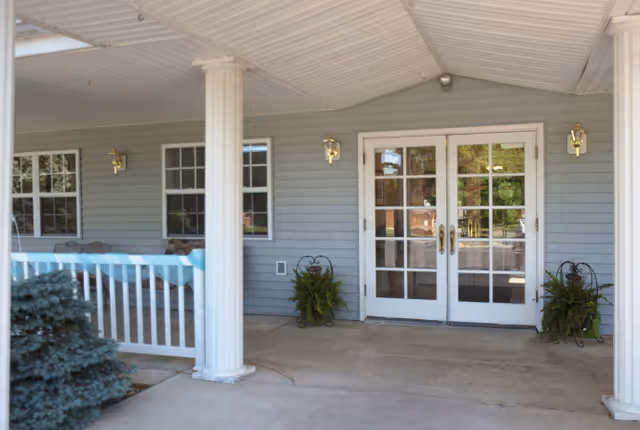 Covered entrance area of a building with white double doors featuring glass panes, flanked by two white columns and two wall-mounted lantern-style lights. There are two potted plants on either side of the doors and a white railing to the left with some greenery visible.