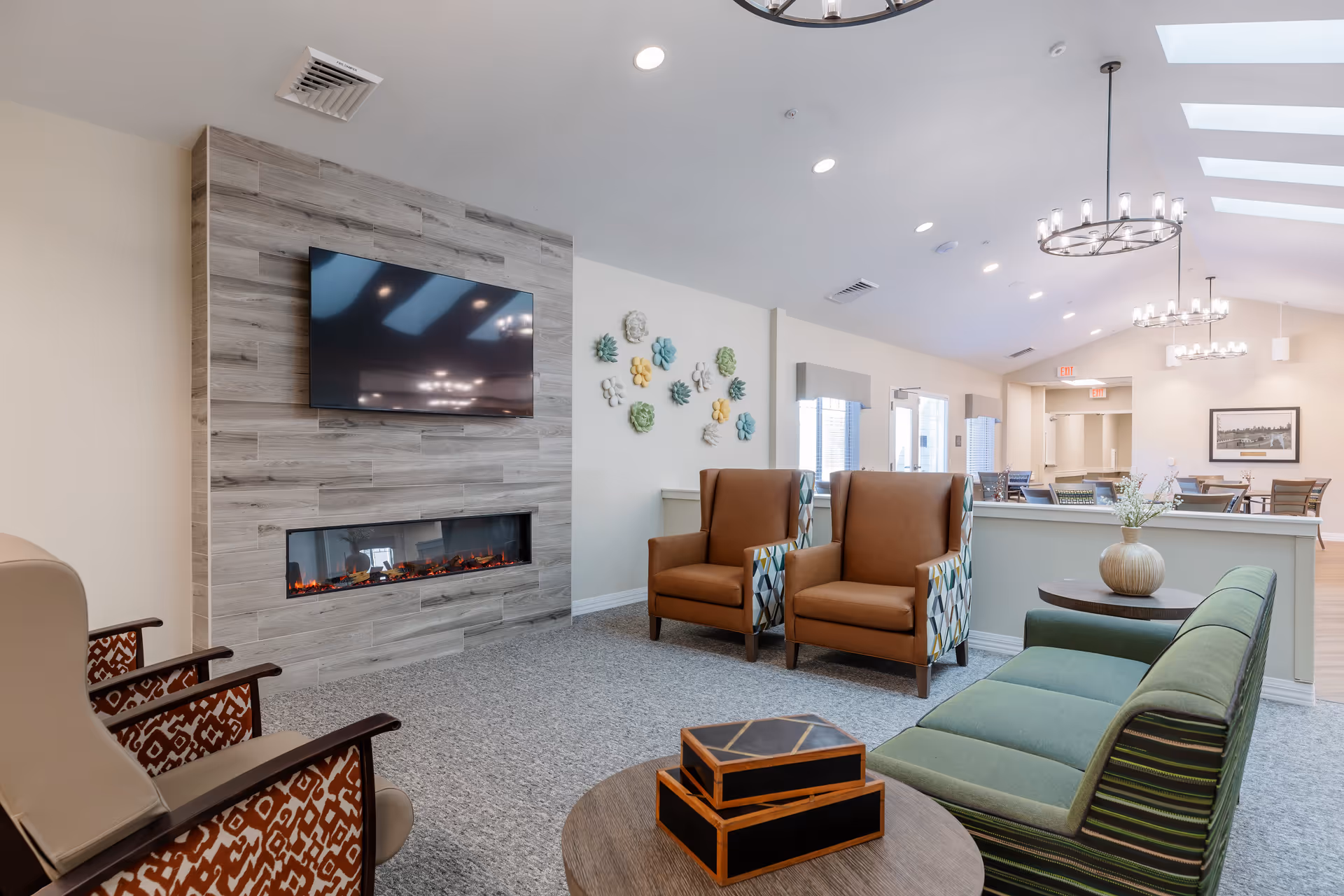 A bright and modern living room area in a senior living facility with a wall-mounted flat-screen TV above a sleek electric fireplace. The room features comfortable seating including two brown armchairs with patterned sides, a green sofa, and two beige chairs with patterned backs. Decorative wall art with colorful flowers is displayed on the wall, and round chandeliers hang from the ceiling with skylights allowing natural light. In the background, there is a dining area with tables and chairs.