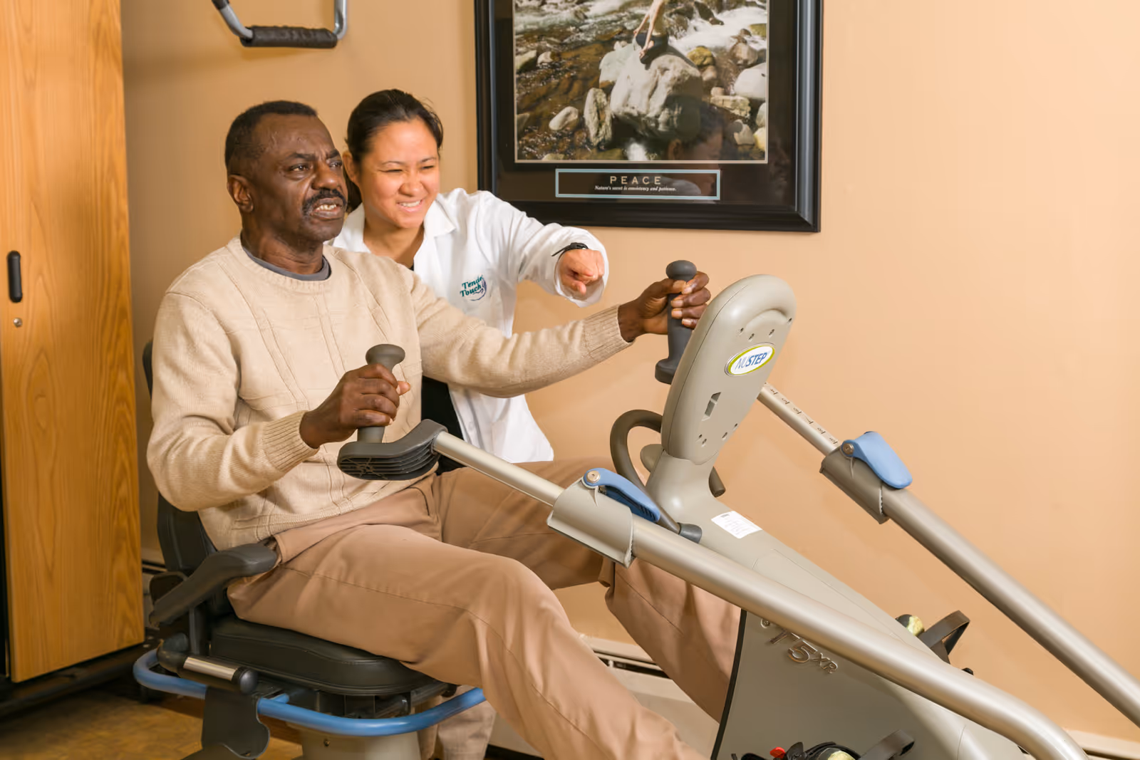 An elderly man is seated on a NuStep exercise machine, holding the handles and exercising his legs. A healthcare professional in a white coat is standing beside him, smiling and pointing forward, providing encouragement. The setting appears to be a therapy or rehabilitation room with a framed picture on the wall and wooden furniture.