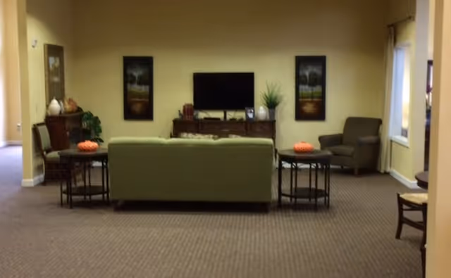 Communal living room with a green sofa facing a wall-mounted TV, side tables, armchairs, and framed artwork.